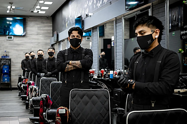 Dos barberos de 2F vestidos de negro y con cubrebocas, de brazos cruzados frente a la hilera de sillones