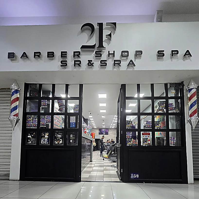 Entrada de 2F Barber Shop Spa en la plaza: pared blanca con el monograma 2F negro y las letras Barber Shop Spa Sr & Sra, mampara negra de cuadrícula abierta al salón y un poste de barbero a cada lado