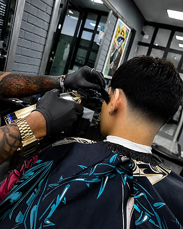 Barbero con guantes negros y reloj dorado delineando la nuca de un cliente sentado de espaldas