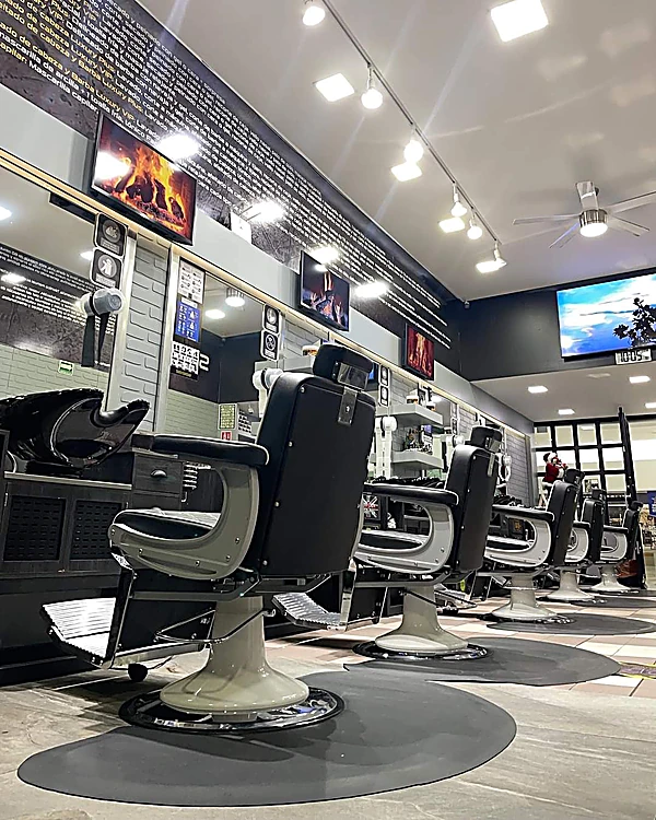 Salón de 2F: fila de sillones de barbero negros, cada uno sobre un tapete redondo negro, piso de ajedrez gris y techo blanco con focos